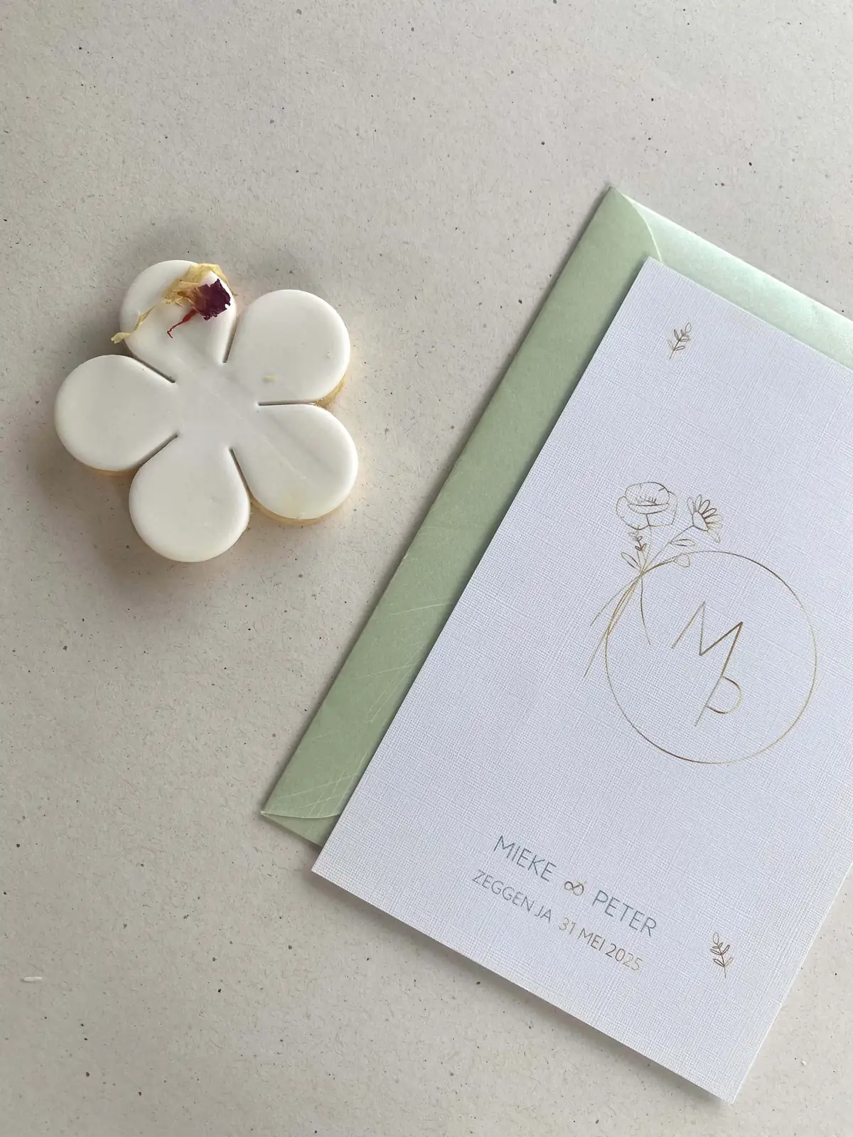 Handgemaakt bloemvormig biscuitje naast een trouwkaart van Mieke en Peter met gouden bloemenmonogram en groene enveloppe