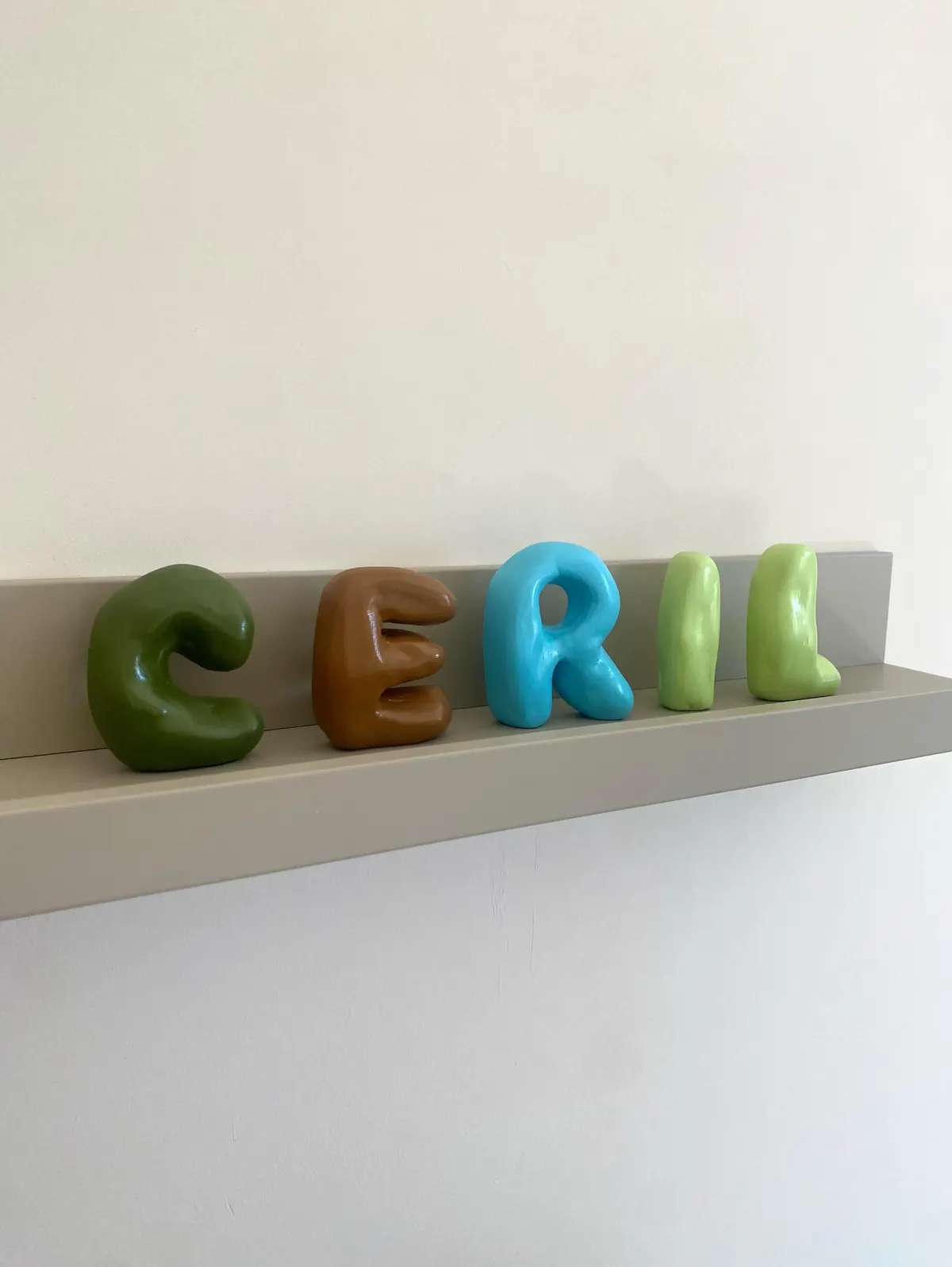 Handgemaakte kleiletters in groen, bruin en blauw die de naam Ceril spellen op een wandplank