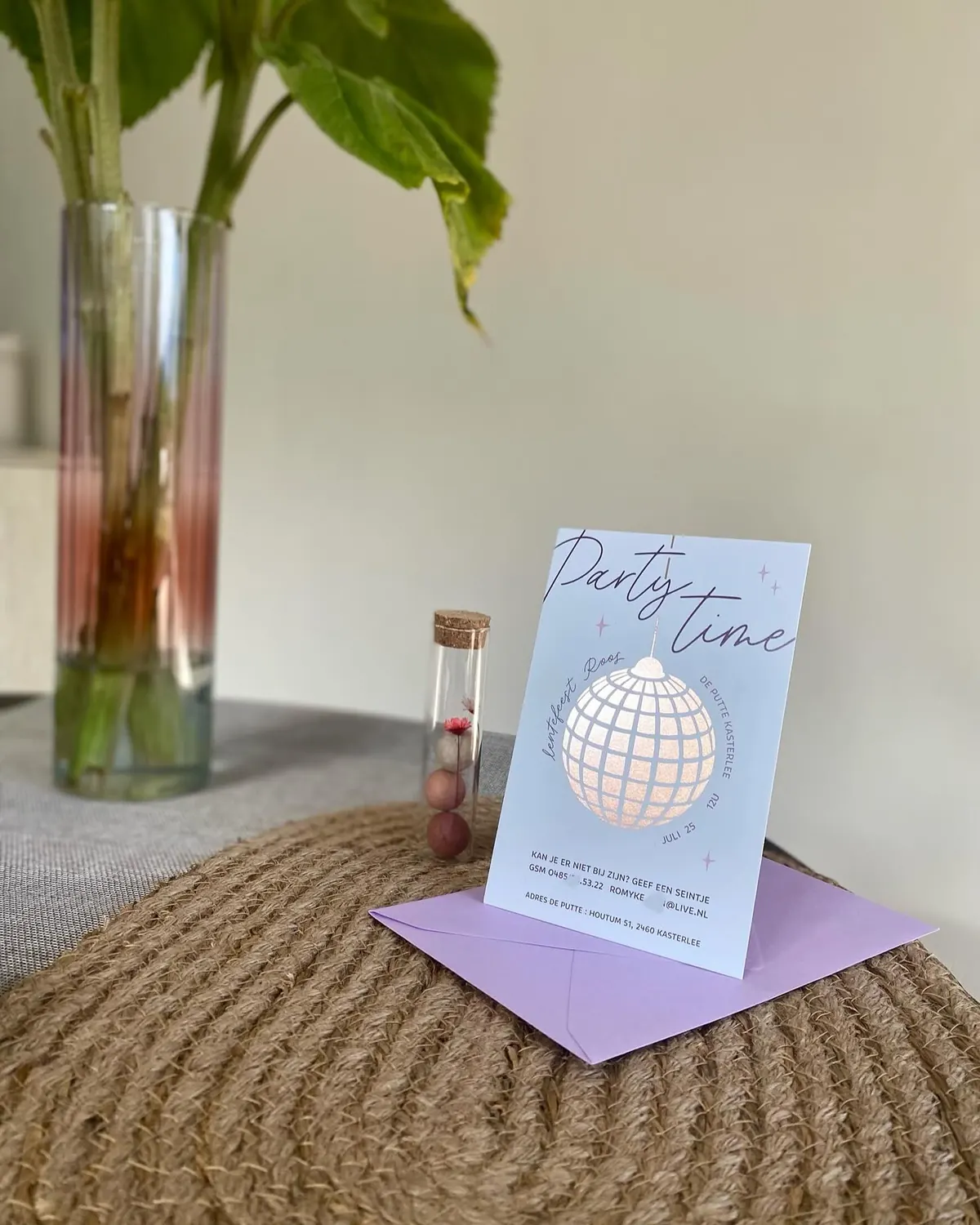 Uitnodiging voor een lentefeest met discobol en de tekst Party time, naast een lila enveloppe en een glazen buisje met droogbloem