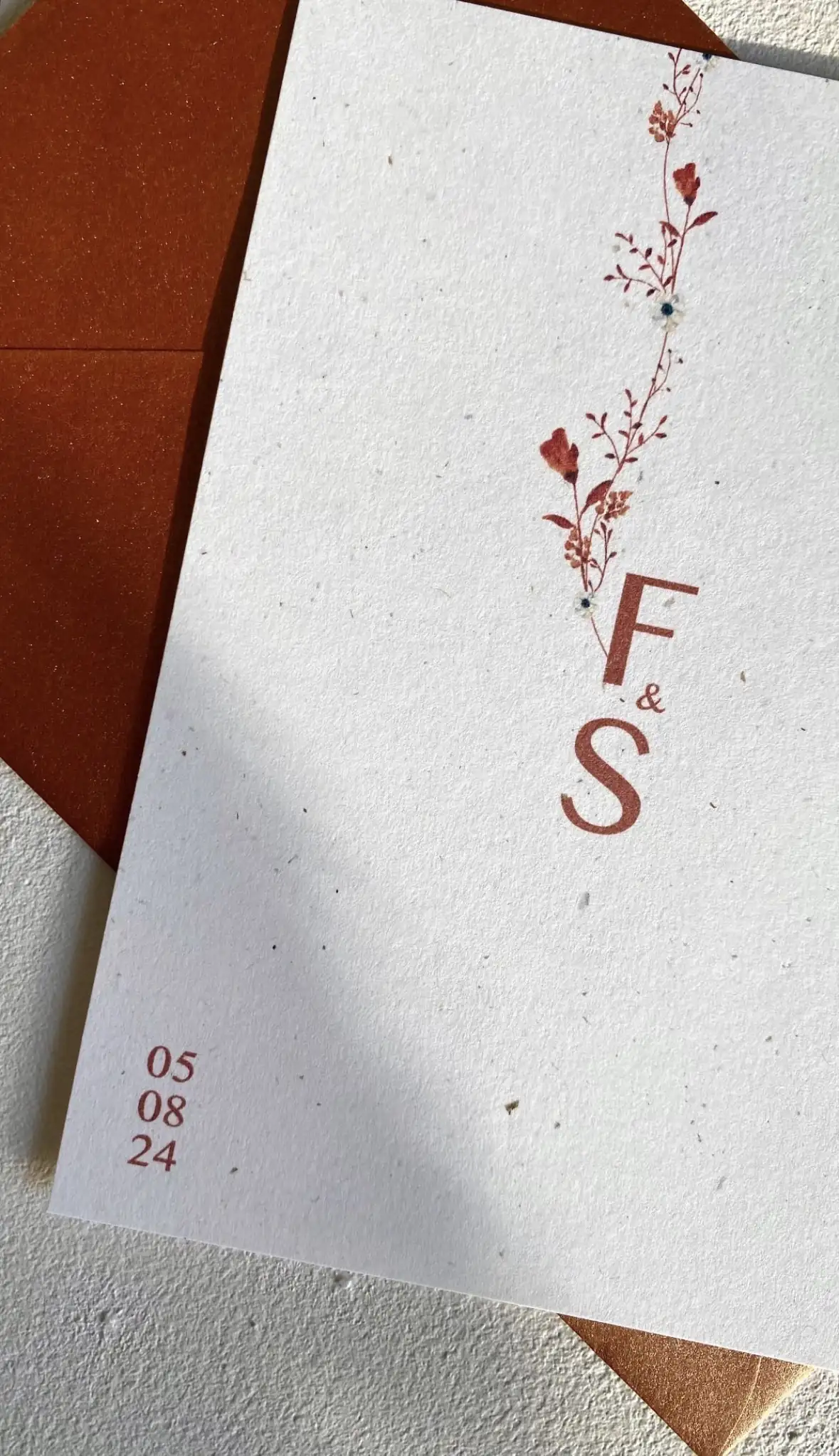 Trouwkaart op maat met bloementak en initialen F & S in terracotta op gespikkeld papier, met koperkleurige enveloppe