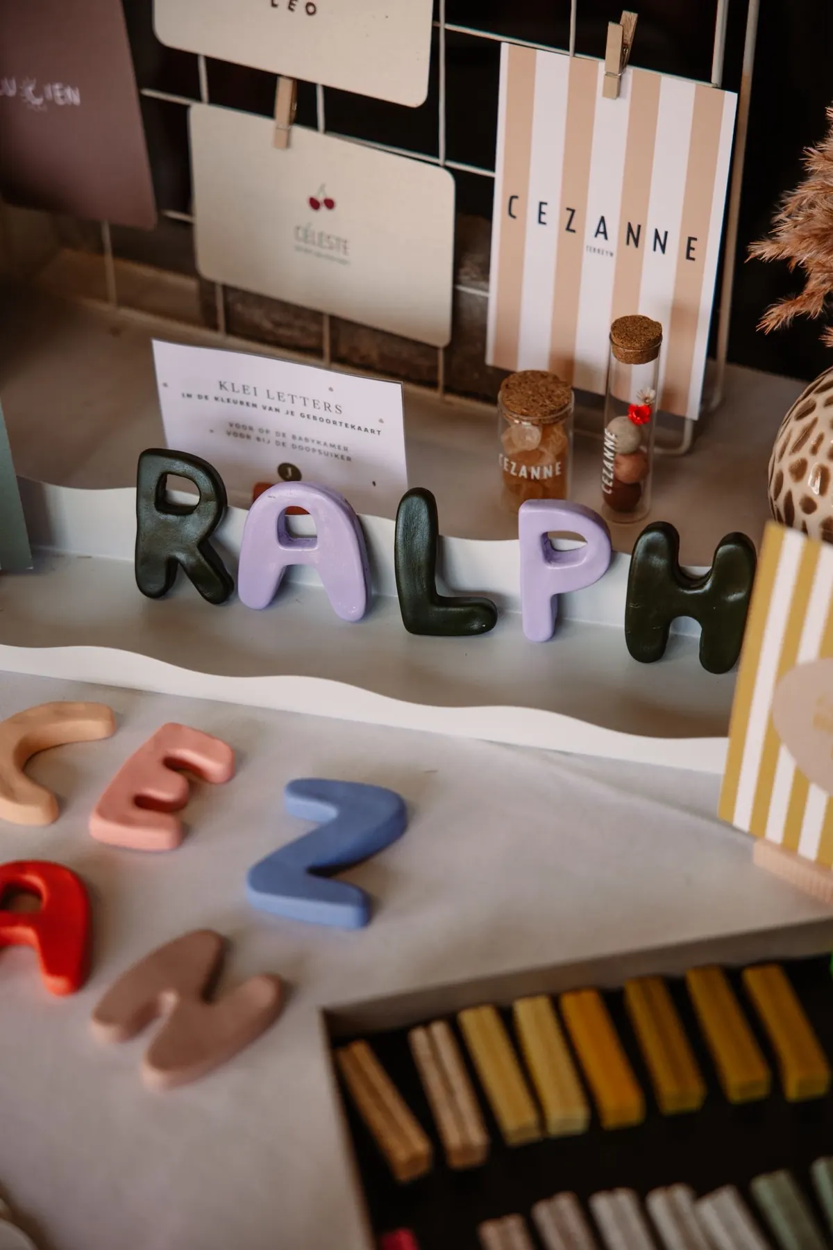 Handgemaakte kleiletters die de naam Ralph spellen, naast geboortekaartjes en doopsuiker